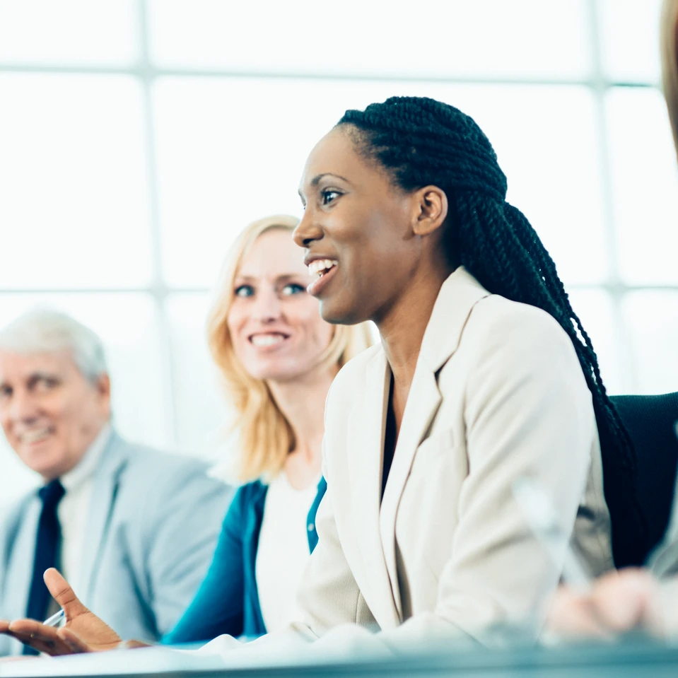 Groupe de professionnels en réunion dans un environnement lumineux. Une femme souriante en costume clair prend la parole, entourée de collègues attentifs et engagés.
